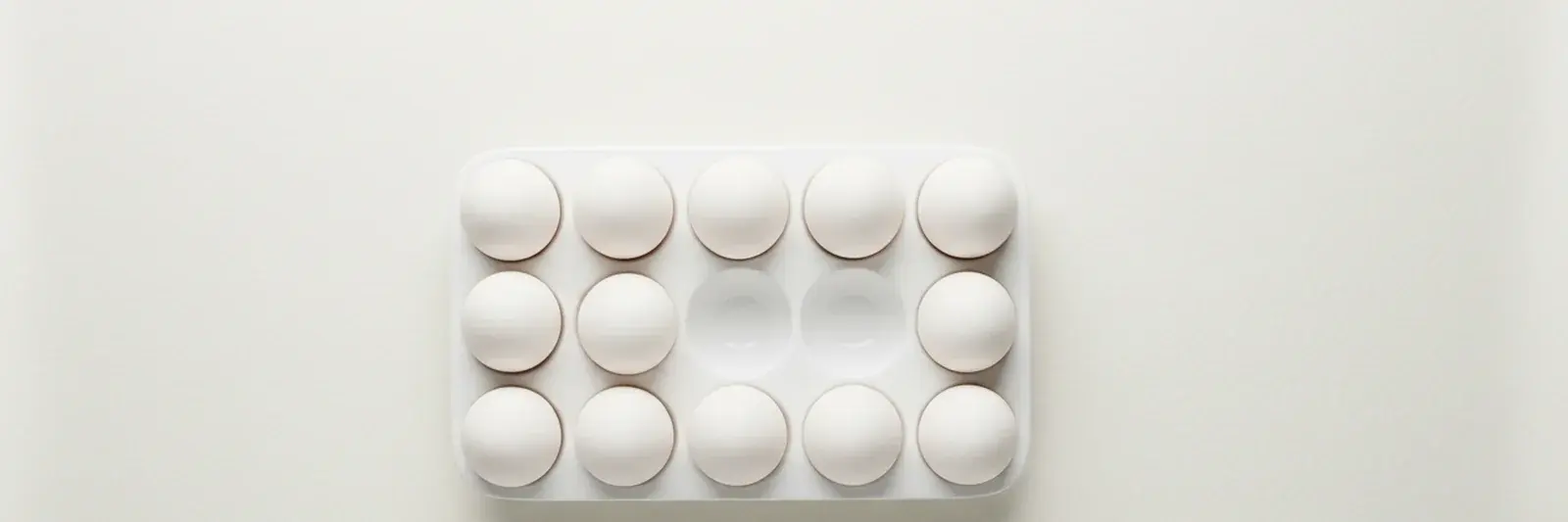 A white ceramic egg tray seen from above on a white surface, eggs in a neat grid with two hollows in the middle left empty.