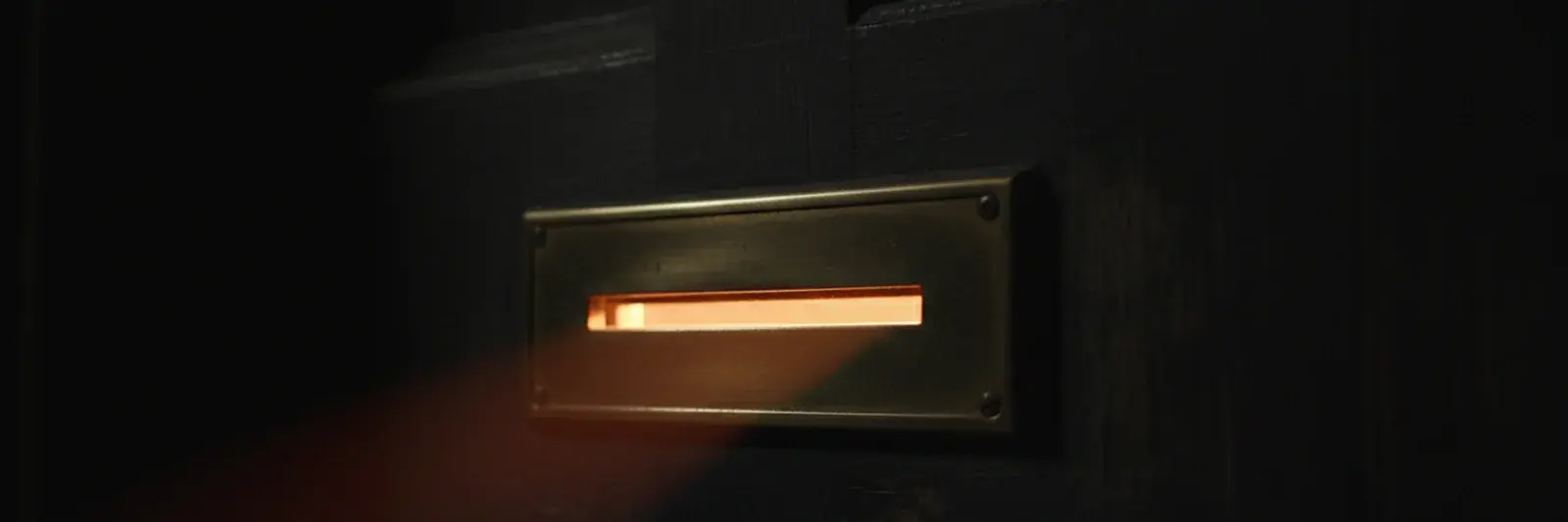 A brass letterbox slot in a dark front door at night, its flap pushed slightly open with warm light from inside spilling through the gap.